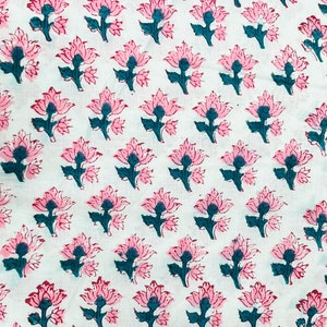 Op de afbeelding: Wit textiel met een herhalend bloemenpatroon. De bloemen zijn roze met donkergroene centra en stelen. Het ontwerp is in blokdrukstijl, wat een textuur geeft. De stof is waarschijnlijk katoen of een vergelijkbaar materiaal.