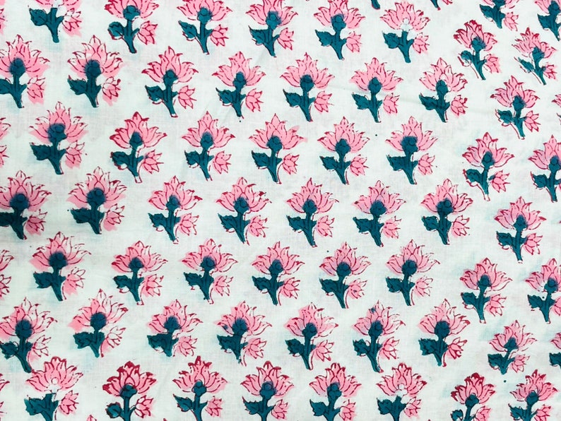 Op de afbeelding: Wit textiel met een herhalend bloemenpatroon. De bloemen zijn roze met donkergroene centra en stelen. Het ontwerp is in blokdrukstijl, wat een textuur geeft. De stof is waarschijnlijk katoen of een vergelijkbaar materiaal.