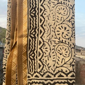 May include: A cream-colored cotton fabric with a black floral and geometric print. The fabric has a gold border with a diamond pattern.