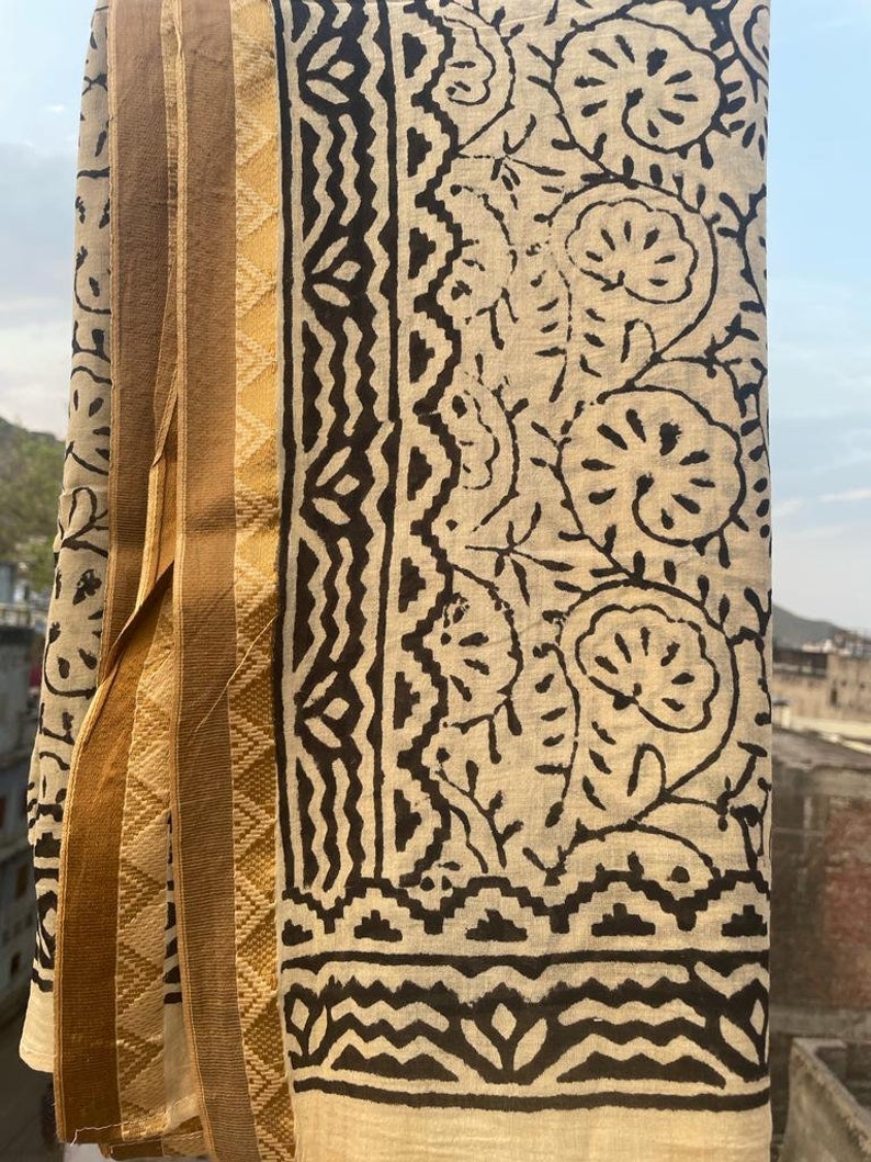 May include: A cream-colored cotton fabric with a black floral and geometric print. The fabric has a gold border with a diamond pattern.