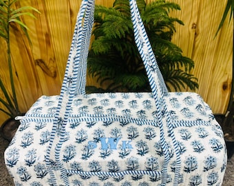 May include: A rectangular quilted duffel bag featuring a white base with a repeating blue floral block print pattern. The bag includes two long striped handles and a central zipper closure. The letters SWK are embroidered in blue on the front.