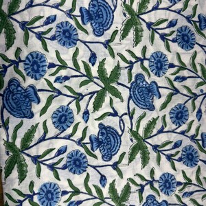 Op de afbeelding: Witte stof met een herhalend patroon van blauwe bloemen en groene bladeren. Het bloemmotief is gedetailleerd en bedekt het gehele oppervlak. De bladeren zijn diepgroen en de bloemen zijn lichtblauw. De achtergrond is wit.