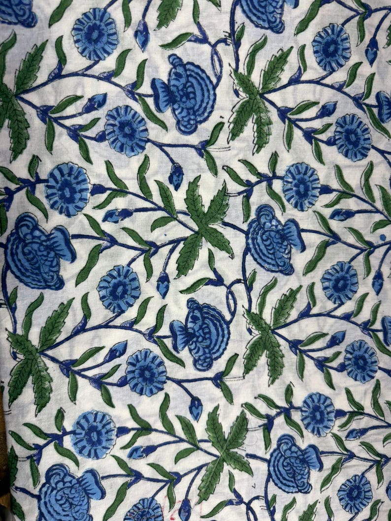 Op de afbeelding: Witte stof met een herhalend patroon van blauwe bloemen en groene bladeren. Het bloemmotief is gedetailleerd en bedekt het gehele oppervlak. De bladeren zijn diepgroen en de bloemen zijn lichtblauw. De achtergrond is wit.