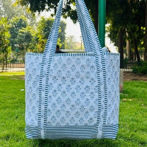 May include: A white tote bag with blue and white striped trim and a blue floral pattern. The bag has two long straps.