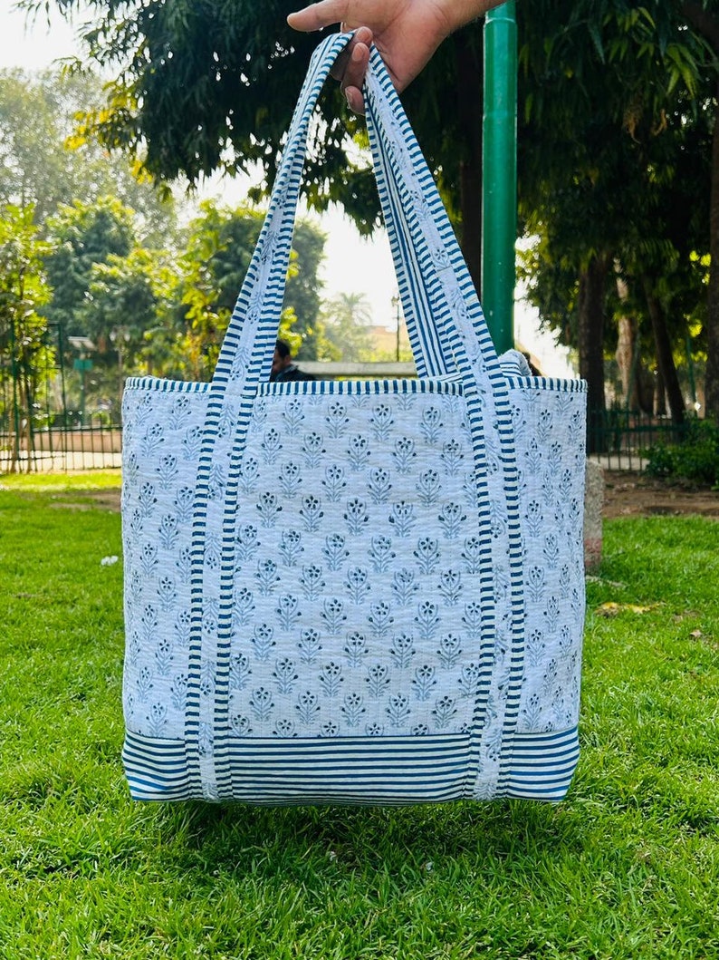 May include: A white tote bag with blue and white striped trim and a blue floral pattern. The bag has two long straps.