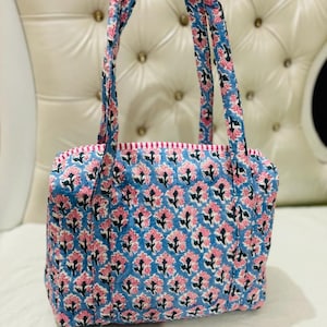 Puede incluir: Una bolsa de mano de algodón azul y rosa con estampado floral y asas dobles. La bolsa está hecha de tela de algodón y tiene un cierre de cremallera.