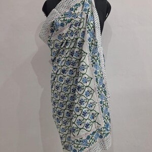 May include: A white cotton fabric wrap featuring a repeating pattern of blue and green floral motifs arranged within a grid of green vines. The edges are finished with a border of small black dots on a white background, creating a distinct frame.