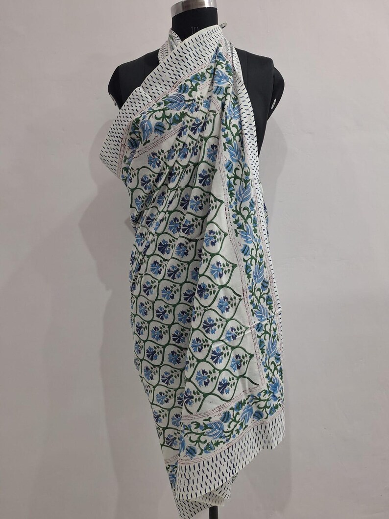 May include: A white cotton fabric wrap featuring a repeating pattern of blue and green floral motifs arranged within a grid of green vines. The edges are finished with a border of small black dots on a white background, creating a distinct frame.