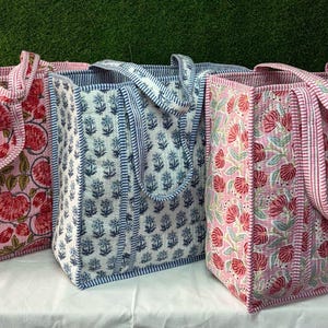 Könnte beinhalten: Drei Tragetaschen mit floralen Mustern. Die Taschen haben eine rechteckige Form mit Griffen. Die Tasche links hat einen rosa Hintergrund mit roten und grünen floralen Designs. Die mittlere Tasche hat einen weißen Hintergrund mit blauen floralen Designs. Die Tasche rechts hat einen rosa Hintergrund mit roten und grünen floralen Designs.