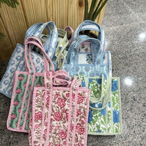 May include: A collection of quilted tote bags with floral and geometric patterns. The bags feature various colors, including pink, blue, and green, with striped handles and borders. The bags are made of fabric and are designed for carrying items.