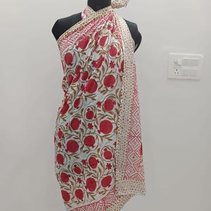 May include: A sarong featuring a white base with a repeating pattern of red pomegranates and green leaves. The fabric includes a border with a tan and white geometric design and a dotted pattern, tied at the neck over a black mannequin torso.
