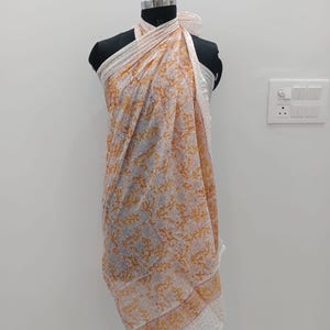 May include: A lightweight sarong featuring a white base with a repeating floral pattern in shades of orange and light blue. The fabric is draped around a mannequin, showcasing a border with a dotted design along the bottom edge and sides.