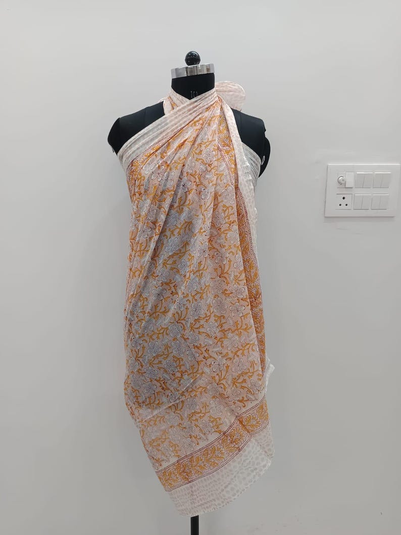 May include: A lightweight sarong featuring a white base with a repeating floral pattern in shades of orange and light blue. The fabric is draped around a mannequin, showcasing a border with a dotted design along the bottom edge and sides.