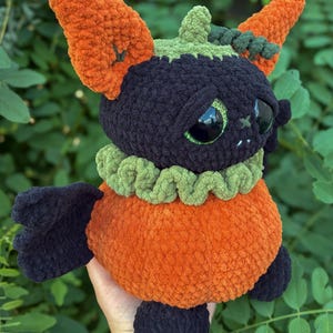 May include: A plush toy shaped like a creature with a black head, large green eyes, and orange ears. It features a green ruffled collar, an orange body, and black wings and feet. The texture is soft and fuzzy, crafted from thick yarn in a crochet style.
