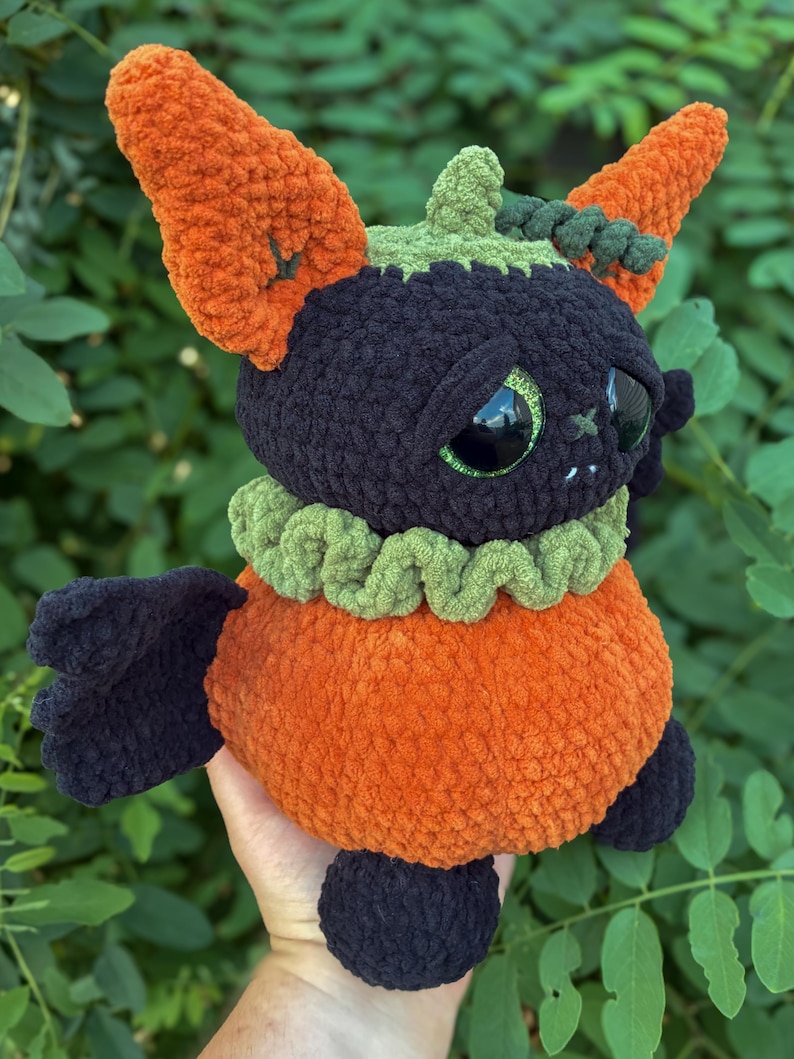 May include: A plush toy shaped like a creature with a black head, large green eyes, and orange ears. It features a green ruffled collar, an orange body, and black wings and feet. The texture is soft and fuzzy, crafted from thick yarn in a crochet style.