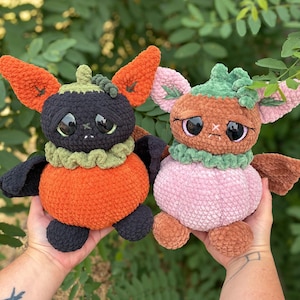 May include: Two handmade crochet plush toys shaped like creatures with pumpkin-like bodies. The left plush has a black head, orange body, and large ears. The right plush has a light pink head, light pink body, and green accents on the head.