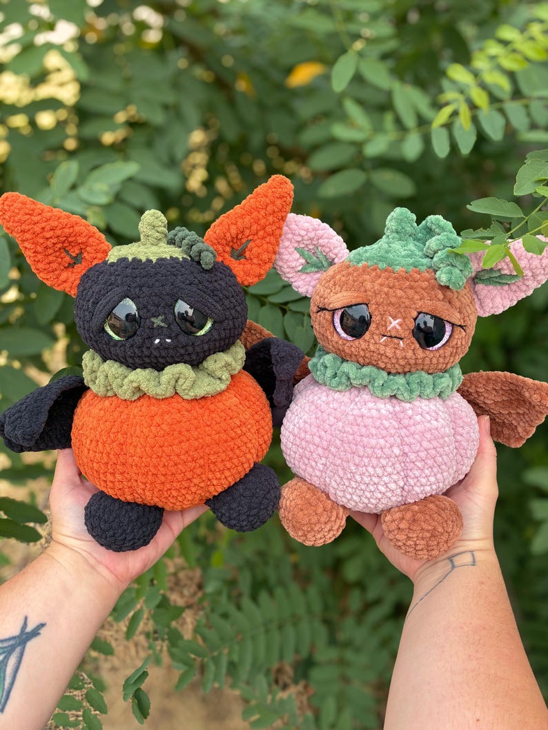 May include: Two handmade crochet plush toys shaped like creatures with pumpkin-like bodies. The left plush has a black head, orange body, and large ears. The right plush has a light pink head, light pink body, and green accents on the head.