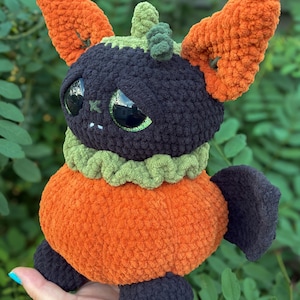 May include: A plush crochet creature featuring a round orange body, a dark purple head with large green eyes, and tall orange ears. It has a green ruffled collar, a small green stem on top of its head, and dark purple feet and wings.