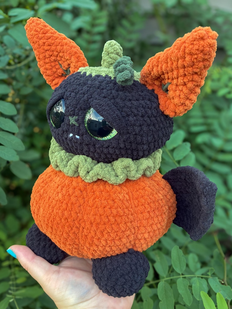 May include: A plush crochet creature featuring a round orange body, a dark purple head with large green eyes, and tall orange ears. It has a green ruffled collar, a small green stem on top of its head, and dark purple feet and wings.