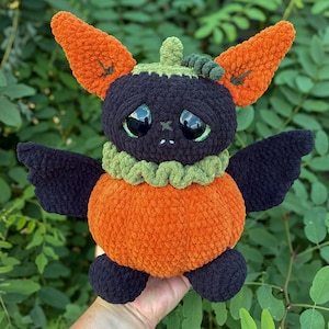 May include: A plush toy shaped like a creature with a round orange pumpkin body, dark purple wings, and large black eyes. It features tall orange ears, a green ruffled collar, and a small green stem on top of its dark purple head.