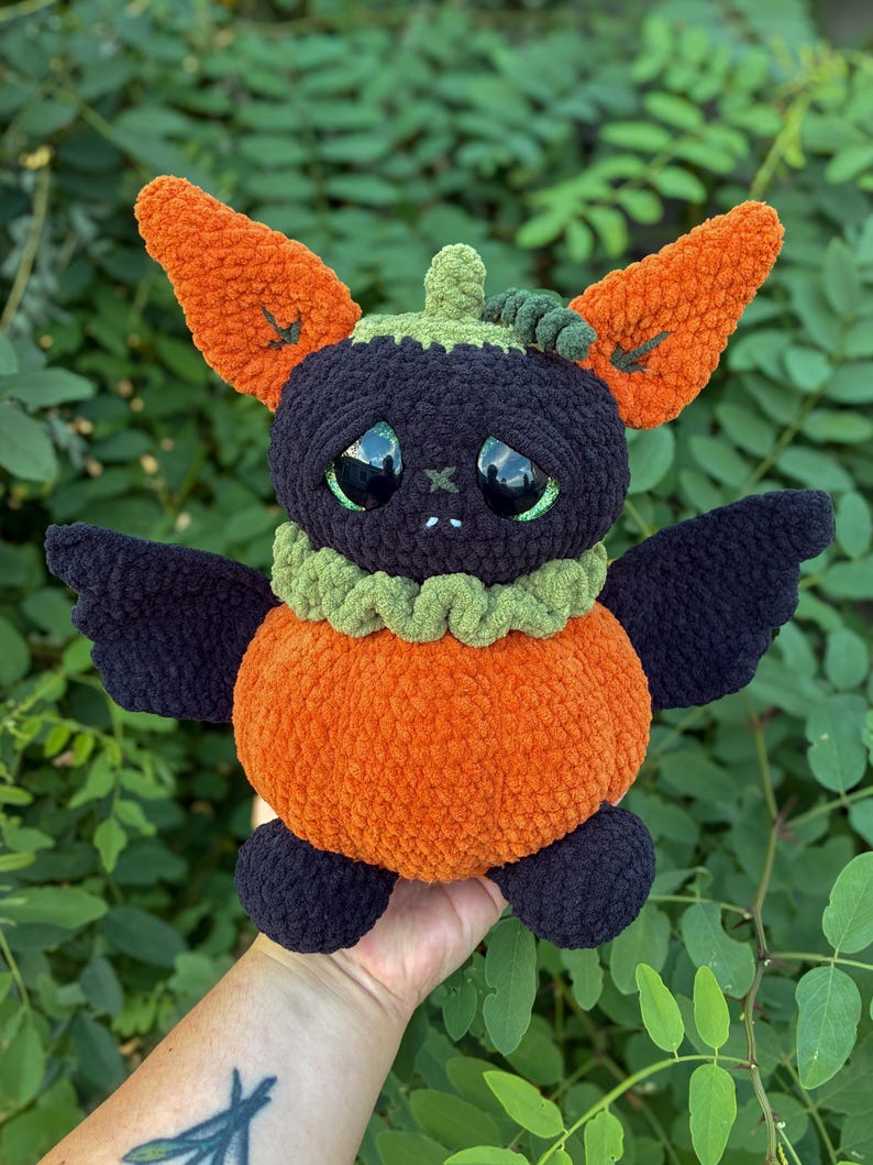 May include: A plush toy shaped like a creature with a round orange pumpkin body, dark purple wings, and large black eyes. It features tall orange ears, a green ruffled collar, and a small green stem on top of its dark purple head.