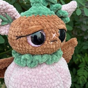May include: A plush toy crafted from soft brown, light pink, and green yarn. The creature features large, round, black glittery eyes, a green ruffled collar, and a green stem-like top. It has light pink ears and a rounded light pink body.