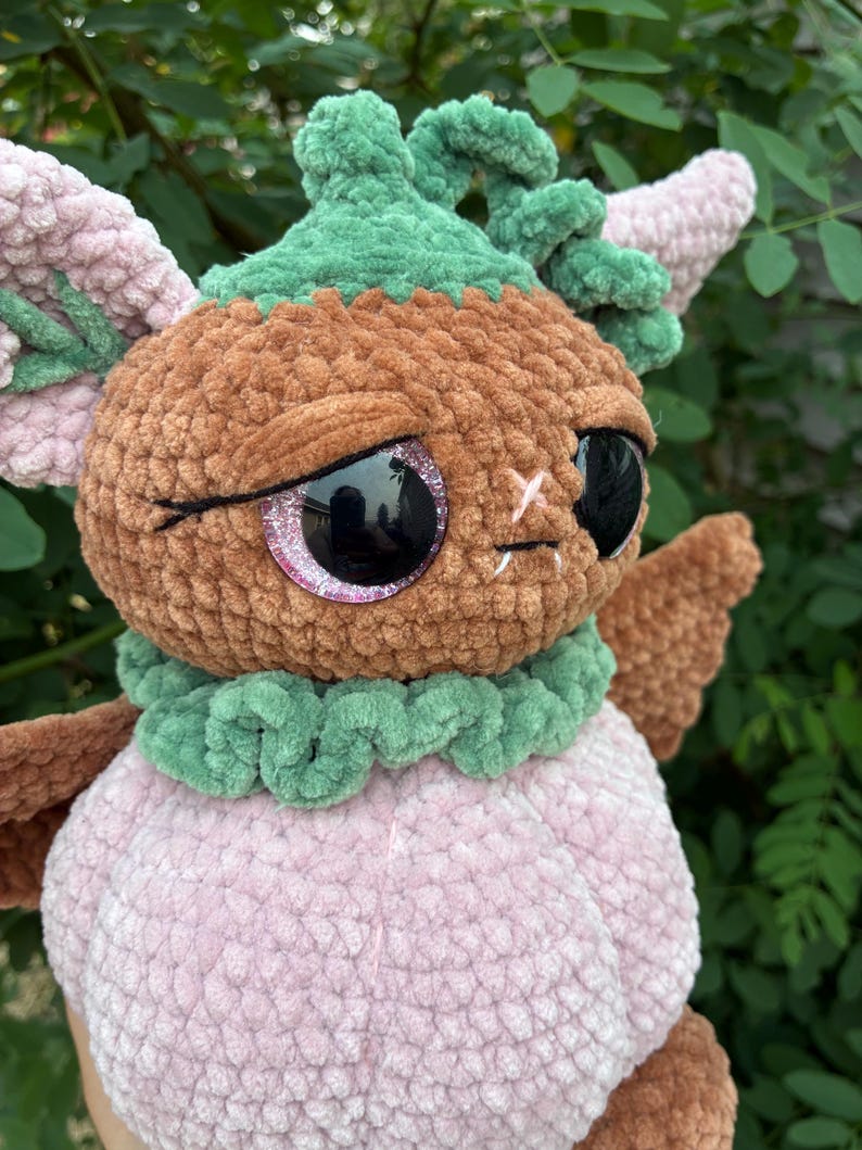 May include: A plush toy crafted from soft brown, light pink, and green yarn. The creature features large, round, black glittery eyes, a green ruffled collar, and a green stem-like top. It has light pink ears and a rounded light pink body.