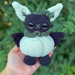 May include: A handmade crochet plush toy featuring a dark purple head with large black eyes and a small green cross-stitched nose. It has large white textured ears, a dark green ruffled collar, a round white body, and dark purple wings and feet.