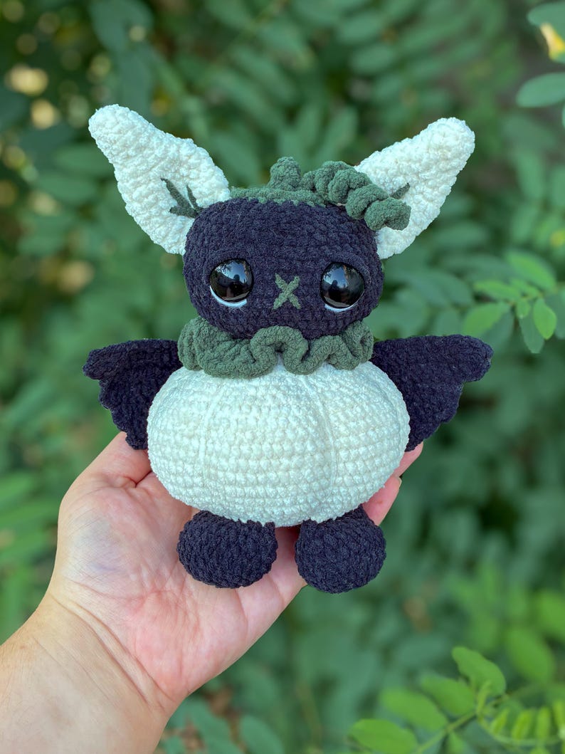 May include: A handmade crochet plush toy featuring a dark purple head with large black eyes and a small green cross-stitched nose. It has large white textured ears, a dark green ruffled collar, a round white body, and dark purple wings and feet.