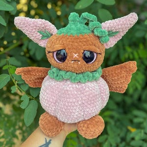 May include: A plush toy creature made of soft yarn with a brown head, large dark eyes, and a light pink pumpkin-shaped body. It features green accents around the neck and top of the head, with pointed pink ears and small brown wings on the sides.