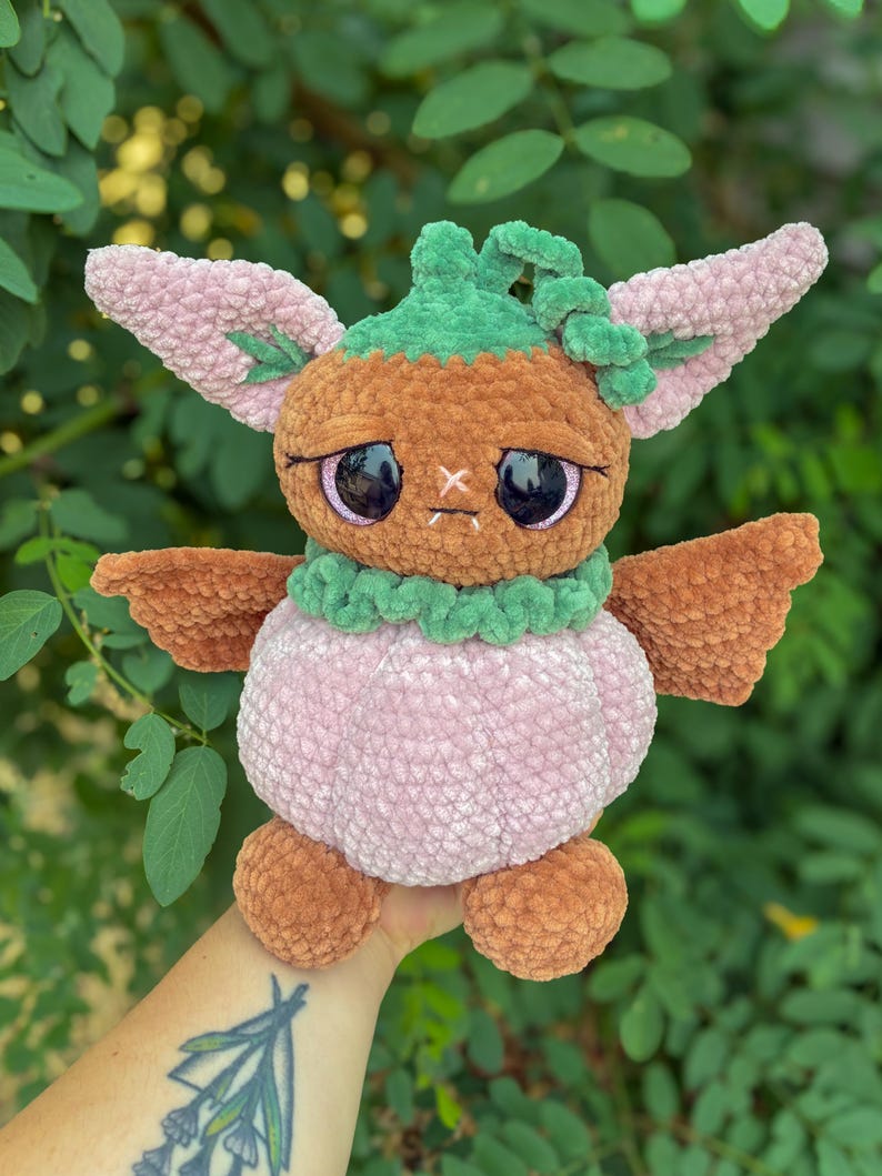 May include: A plush toy creature made of soft yarn with a brown head, large dark eyes, and a light pink pumpkin-shaped body. It features green accents around the neck and top of the head, with pointed pink ears and small brown wings on the sides.