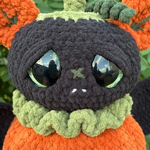 May include: A plush crochet creature featuring a round black head with large reflective green eyes, a small stitched X-shaped nose, and two white fangs. It has a ruffled light green collar, an orange pumpkin-shaped body, and orange ears.