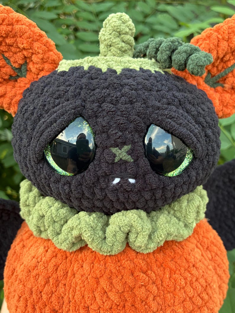 May include: A plush crochet creature featuring a round black head with large reflective green eyes, a small stitched X-shaped nose, and two white fangs. It has a ruffled light green collar, an orange pumpkin-shaped body, and orange ears.