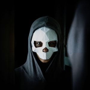 Op de afbeelding: Een persoon draagt een grijze hoodie met een wit origami-schedelmasker.