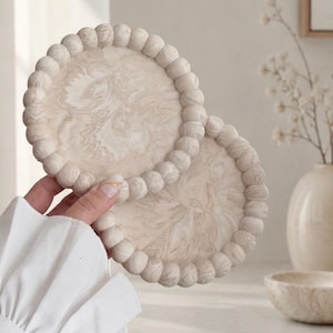 May include: Two round, beige coasters with a marbled design and a beaded edge. The coasters are held in a hand with a white sleeve in the background. The coasters are a decorative home accessory.