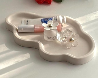 May include: A light beige, cloud-shaped tray holds beauty products, including a lip gloss, a cream tube, and two perfume bottles with daisy-shaped caps. A red rose rests nearby, adding a pop of color to the neutral palette.