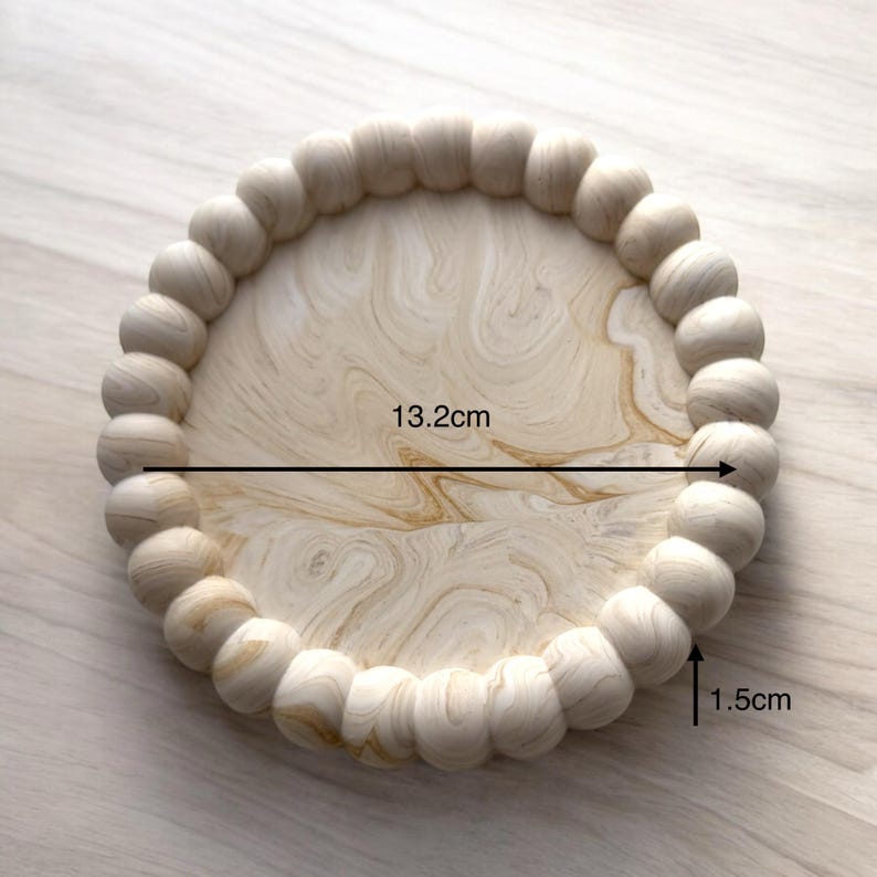 May include: A round wooden tray with a beaded edge. The tray has a light wood grain pattern and measures 13.2 cm in diameter and 1.5 cm in height. The tray is suitable for serving or display.