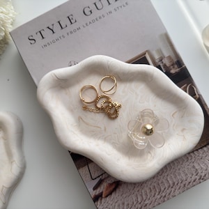 Peut inclure: Un plateau décoratif blanc et crème marbré avec des bijoux en or et un porte-bague transparent en forme de fleur. Le plateau est posé sur un magazine avec le texte "STYLE GUIDE". Les bijoux comprennent des bagues et une chaîne.