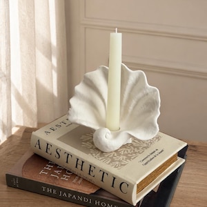 Peut inclure: Un bougeoir blanc en forme de coquillage avec une bougie crème, posé sur deux livres. Les livres sont intitulés "Aesthetic" et "The Japandi Home". Le bougeoir a un motif en spirale à la base.