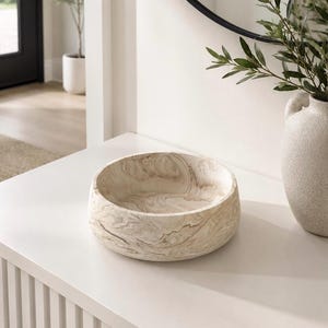 May include: A round, wooden decorative bowl with a light brown and cream marbled pattern. The bowl sits on a white surface, with a white vase and greenery in the background. The bowl is a decorative accent piece.