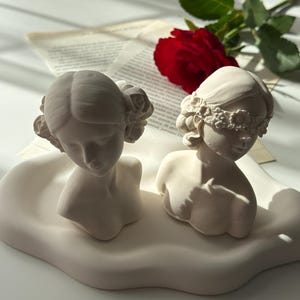 Puede incluir: Dos bustos de mujeres de yeso blanco con diademas florales. Un busto tiene una venda en los ojos. Los bustes est&aacute;n sobre una base blanca en forma de nube.