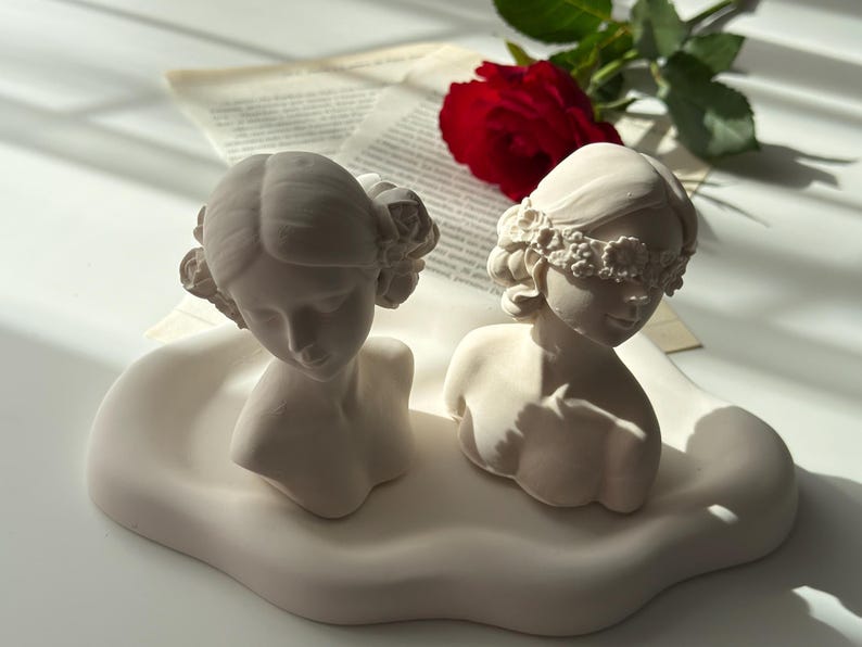 Puede incluir: Dos bustos de mujeres de yeso blanco con diademas florales. Un busto tiene una venda en los ojos. Los bustes est&aacute;n sobre una base blanca en forma de nube.