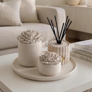 May include: A neutral-toned home decor arrangement featuring a round tray with two lidded jars topped with flower-shaped designs and a reed diffuser with black reeds. The items are arranged on a white coffee table, creating a minimalist aesthetic.
