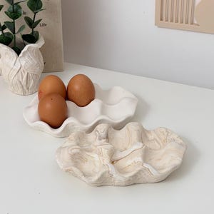 May include: A white ceramic egg holder with three brown eggs, and a decorative marbled ceramic tray. The egg holder has a wavy design, and the tray has a beige and brown swirl pattern. Home decor.