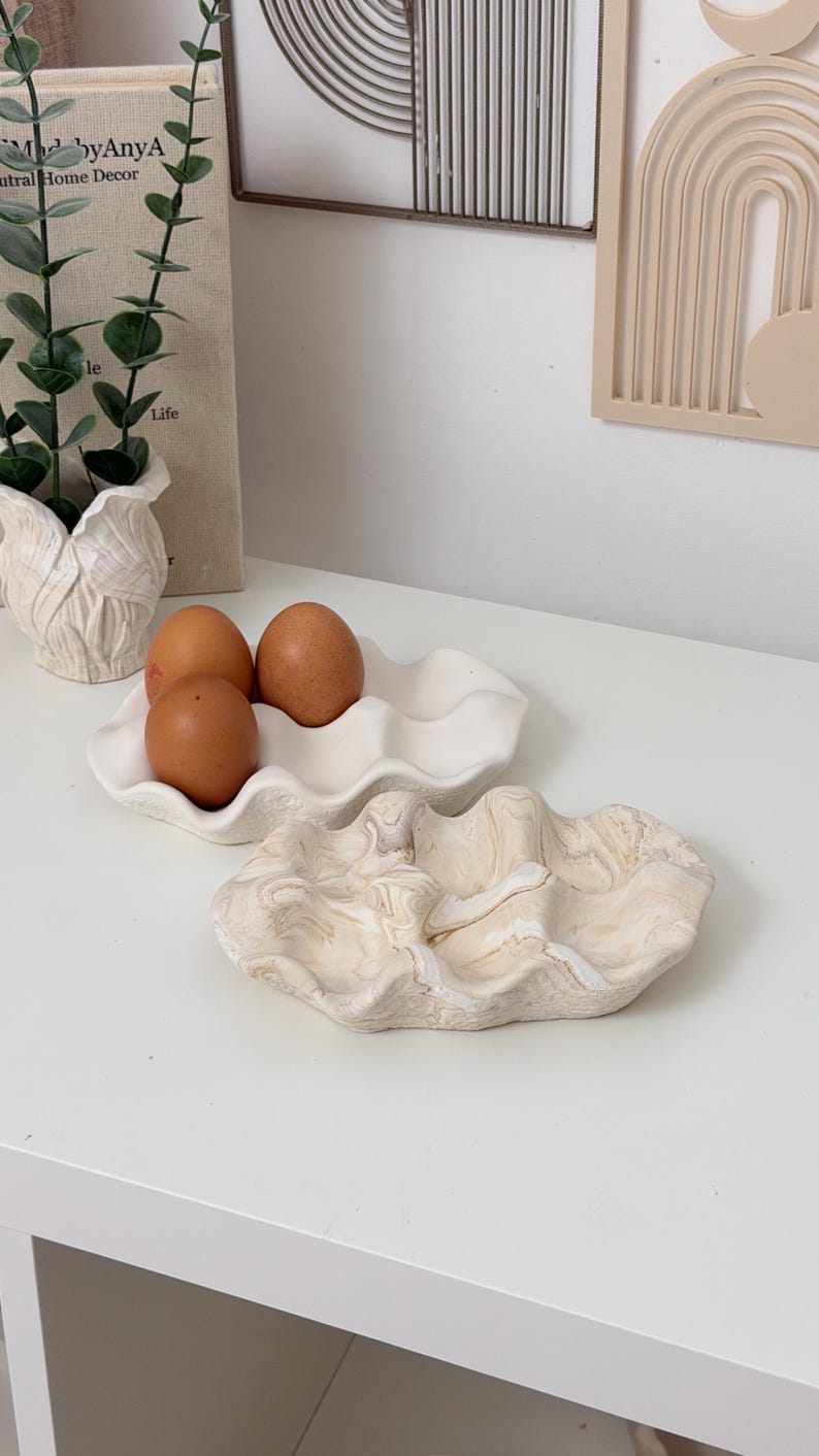 May include: A white ceramic egg holder with three brown eggs, and a decorative marbled ceramic tray. The egg holder has a wavy design, and the tray has a beige and brown swirl pattern. Home decor.