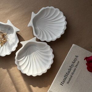 May include: Three white ceramic seashell-shaped dishes. The dishes are on a light brown surface. One dish has a gold chain necklace inside. The text "HandMadebyAnya Neutral Home Decor" is on a book in the background.