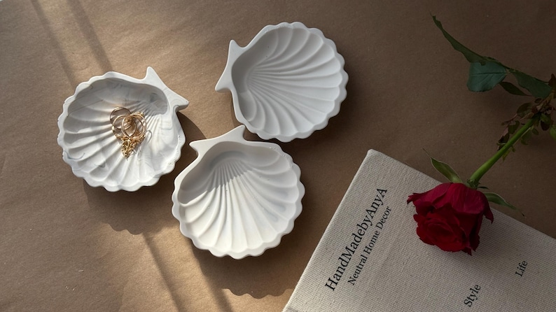 May include: Three white ceramic seashell-shaped dishes. The dishes are on a light brown surface. One dish has a gold chain necklace inside. The text "HandMadebyAnya Neutral Home Decor" is on a book in the background.