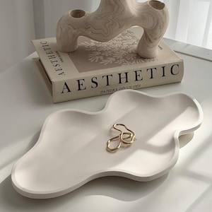 Peut inclure: Composition de décoration d'intérieur esthétique avec un plateau crème de forme abstraite et une bague ondulée dorée. Une sculpture décorative beige est posée sur un livre intitulé "Aesthetic".