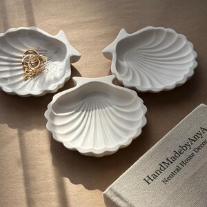 May include: Three white ceramic seashell-shaped dishes. The dishes are arranged in a triangular shape on a beige surface. The dish on the left has a gold chain and rings inside.