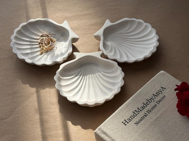 May include: Three white ceramic seashell-shaped dishes. The dishes are arranged in a triangular shape on a beige surface. The dish on the left has a gold chain and rings inside.